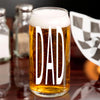 A glass of beer with the word "DAD" printed on it, placed on a table with a checkered flag in the background.