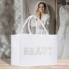 A white tote bag with the word "BRAUT" printed on it, placed on a wooden surface. In the background, there is a woman wearing a white wedding dress, visible in a mirror.