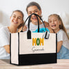 A large tote bag with the text "MOM Savannah" printed on it, placed on a wooden surface. Behind the bag, there are three smiling women, likely a mother and her two daughters.