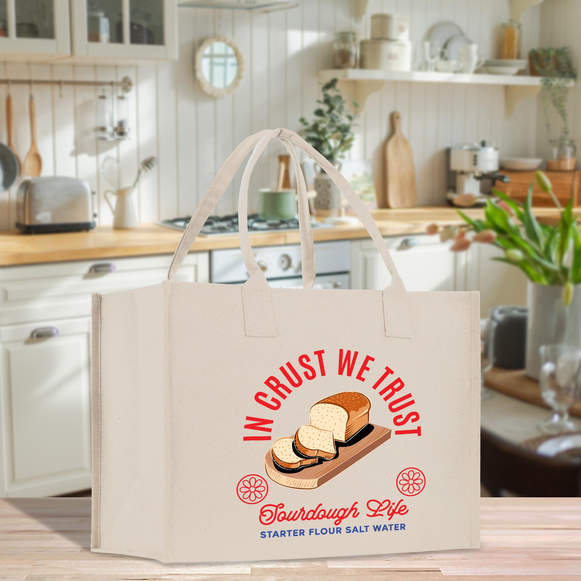 A white tote bag with a graphic design featuring a loaf of bread and the text &quot;InCRUST WE TRUST&quot; and &quot;TOURDOUGH LIFE STARTER FLOUR SALT WATER&quot;.