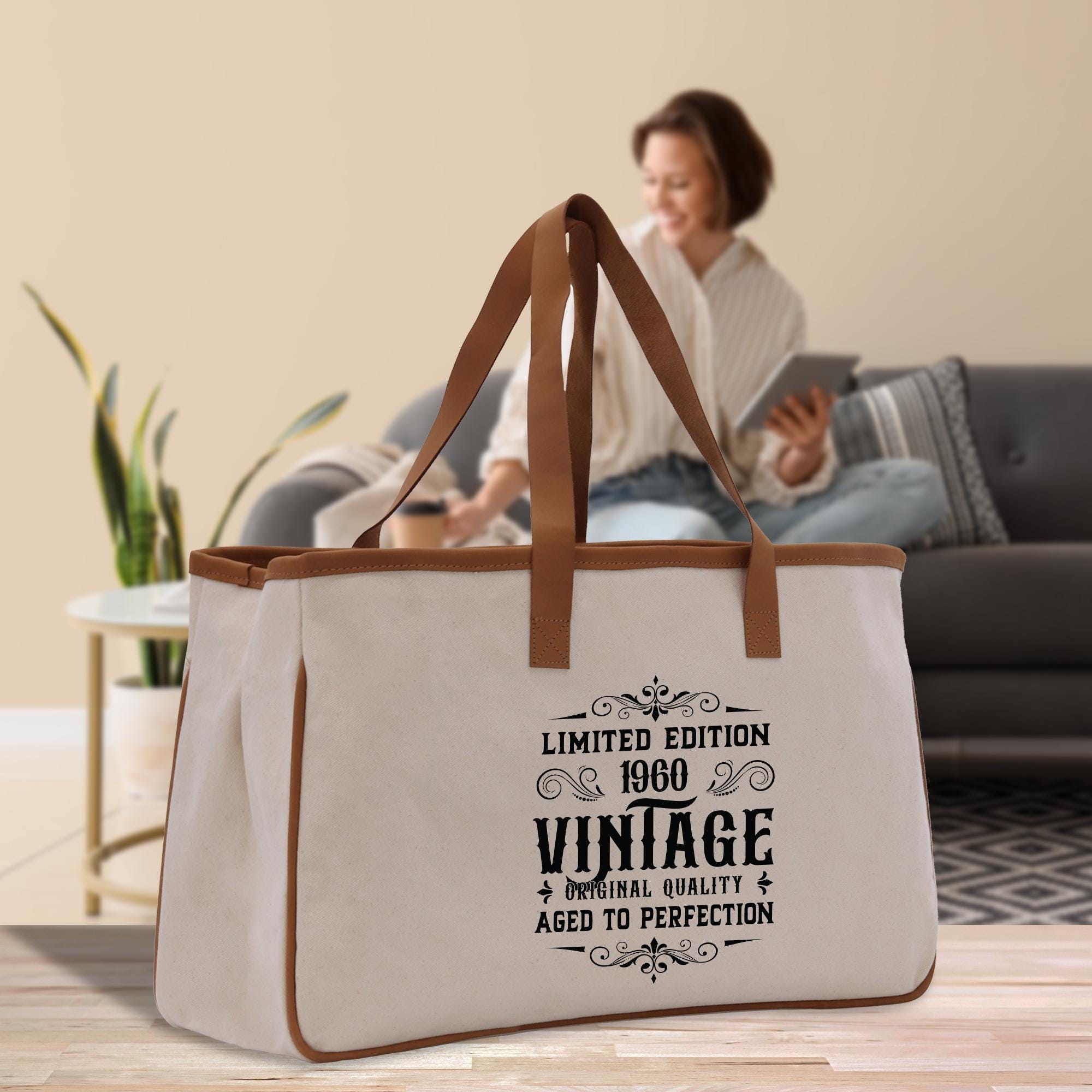 a large white tote bag with a vintage-style design, placed on a wooden floor in a living room setting. A woman is sitting on a couch in the background, and there is a potted plant nearby.