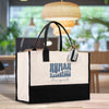 A large white tote bag with black handles and a black and white striped tag is displayed on a wooden surface, with a blurred background that includes plants and a hanging light fixture.