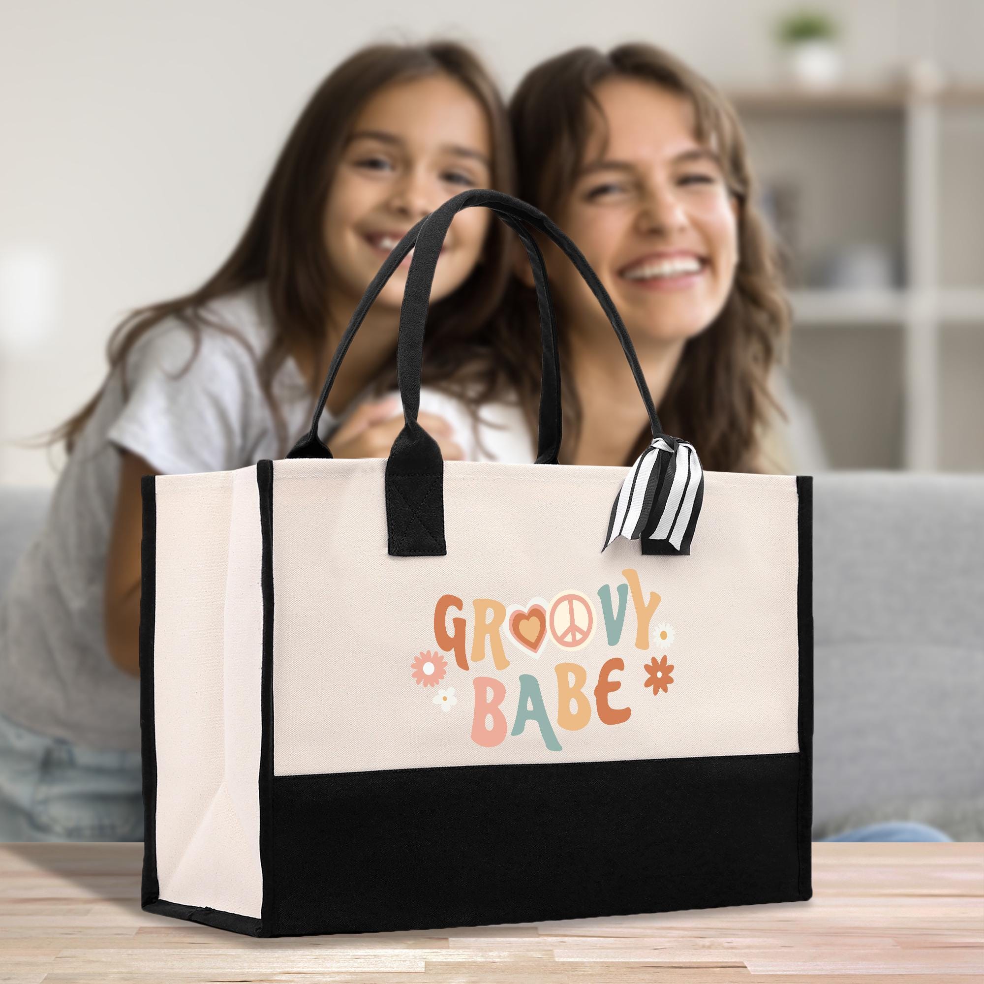 Two young women are smiling and posing behind a large tote bag with the text &quot;GROOVY BABE&quot; printed on it.
