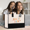 Two young women are smiling and posing behind a large tote bag with the text "GROOVY BABE" printed on it.