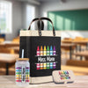 A black tote bag with a handle, a glass jar with a straw, and a small metal container with a lid, all featuring colorful crayon designs, are displayed on a wooden surface.