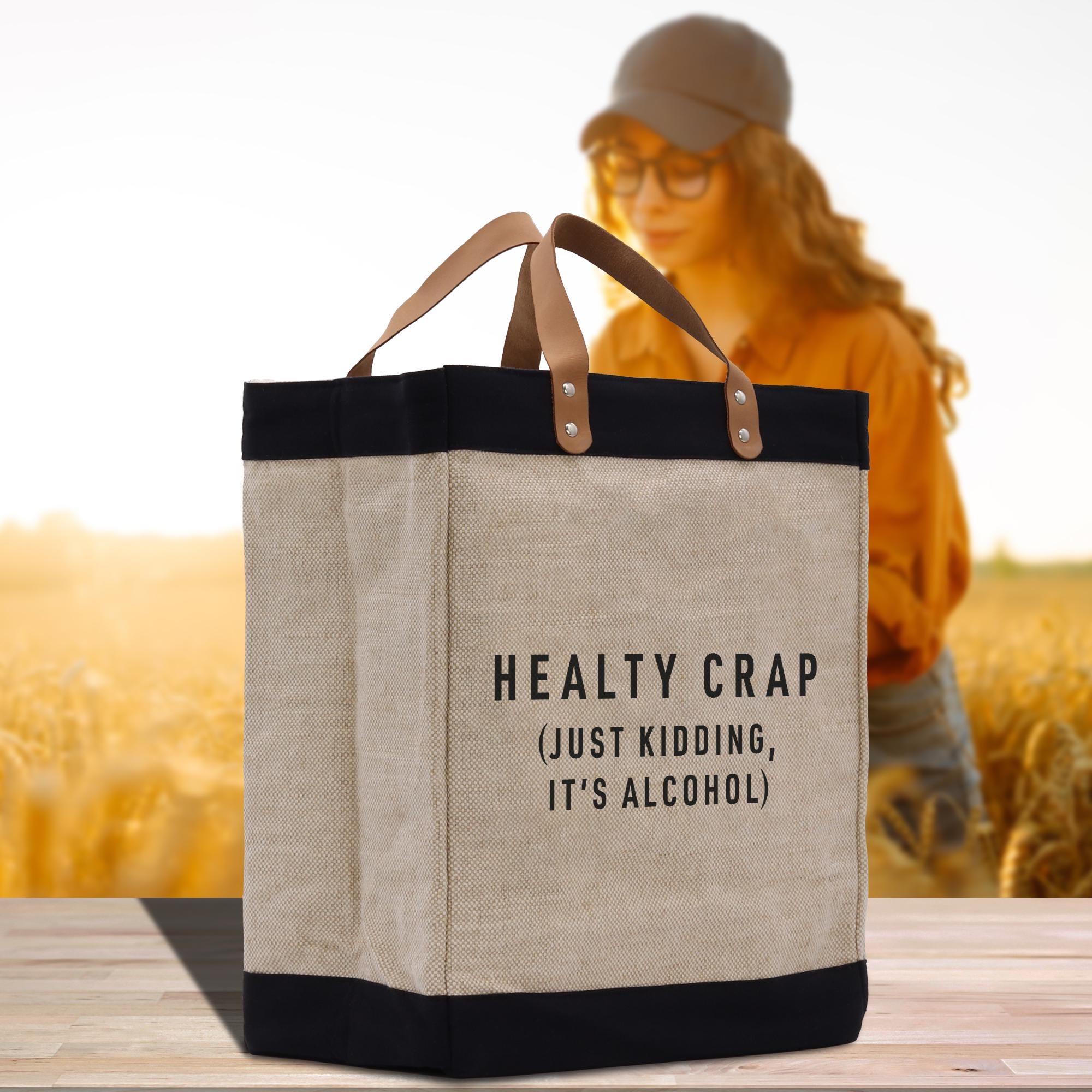 A beige tote bag with black trim and a humorous text printed on it, placed on a wooden surface. In the background, there is a blurred image of a person wearing a hat and glasses.