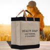 A beige tote bag with black trim and a humorous text printed on it, placed on a wooden surface. In the background, there is a blurred image of a person wearing a hat and glasses.