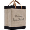 A beige tote bag with black trim and leather handles, featuring personalized text on the front.