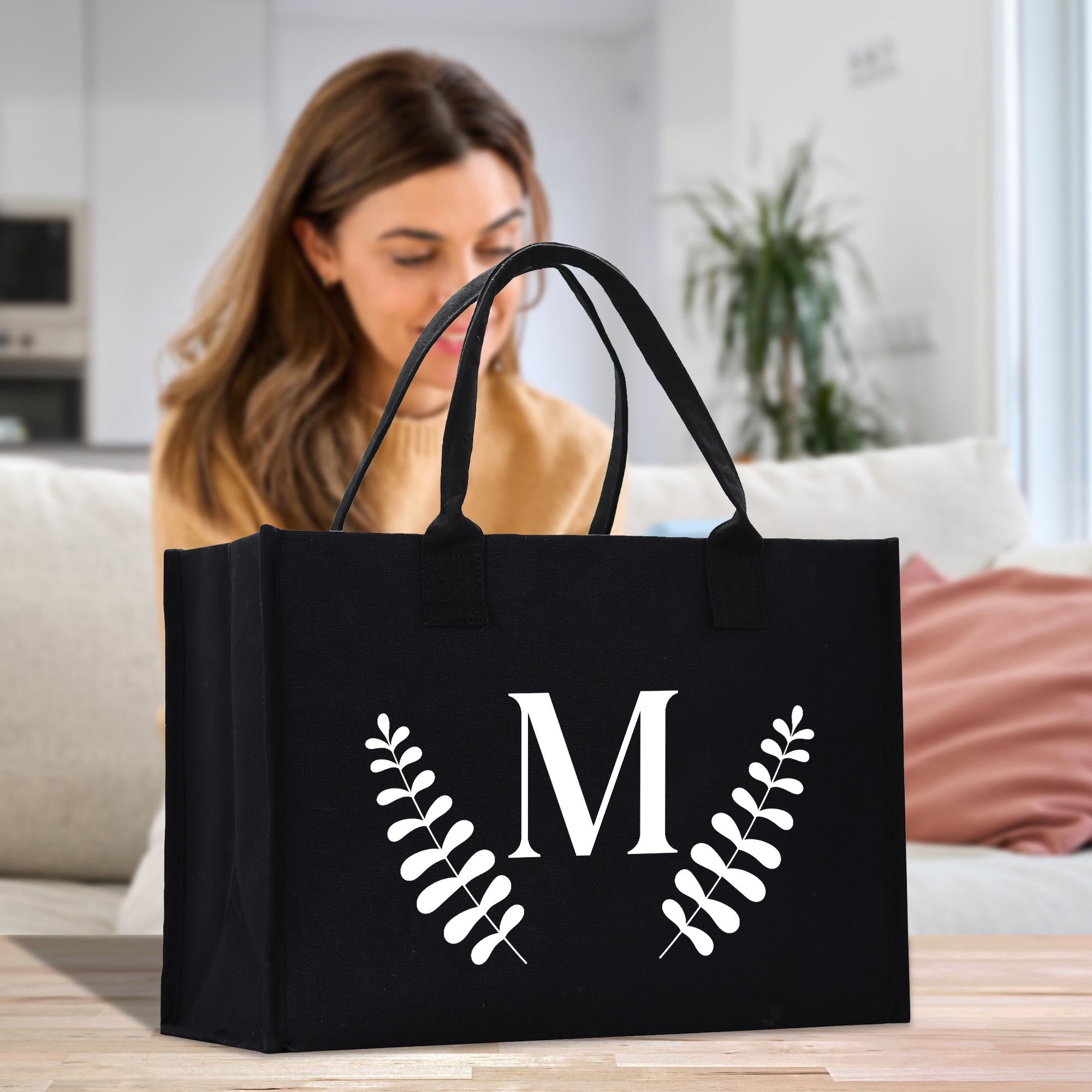 A woman is sitting on a couch in a living room, looking at a black tote bag with a white letter &quot;M&quot; and leaf design on it.