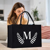 A woman is sitting on a couch in a living room, looking at a black tote bag with a white letter "M" and leaf design on it.
