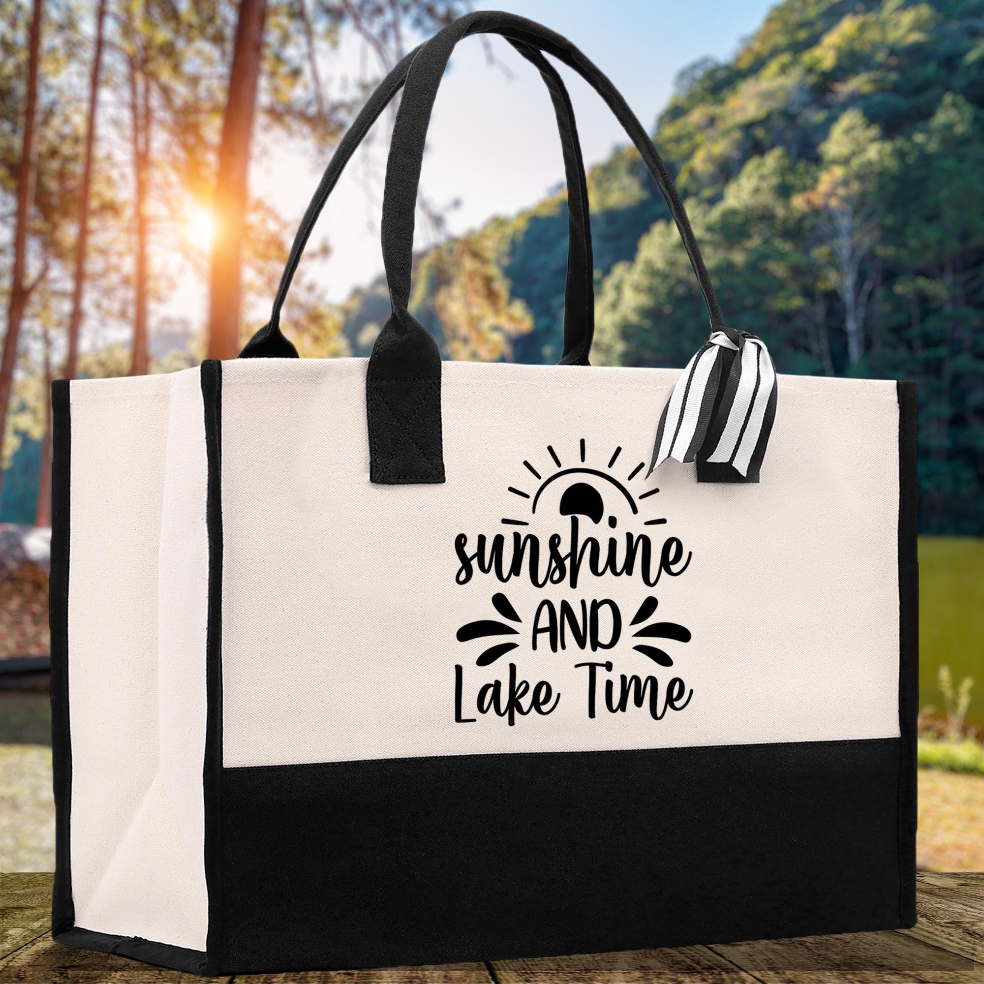 A black and white tote bag with a printed phrase "sunshine and Lake Time" on it, placed on a wooden surface with a natural outdoor background.