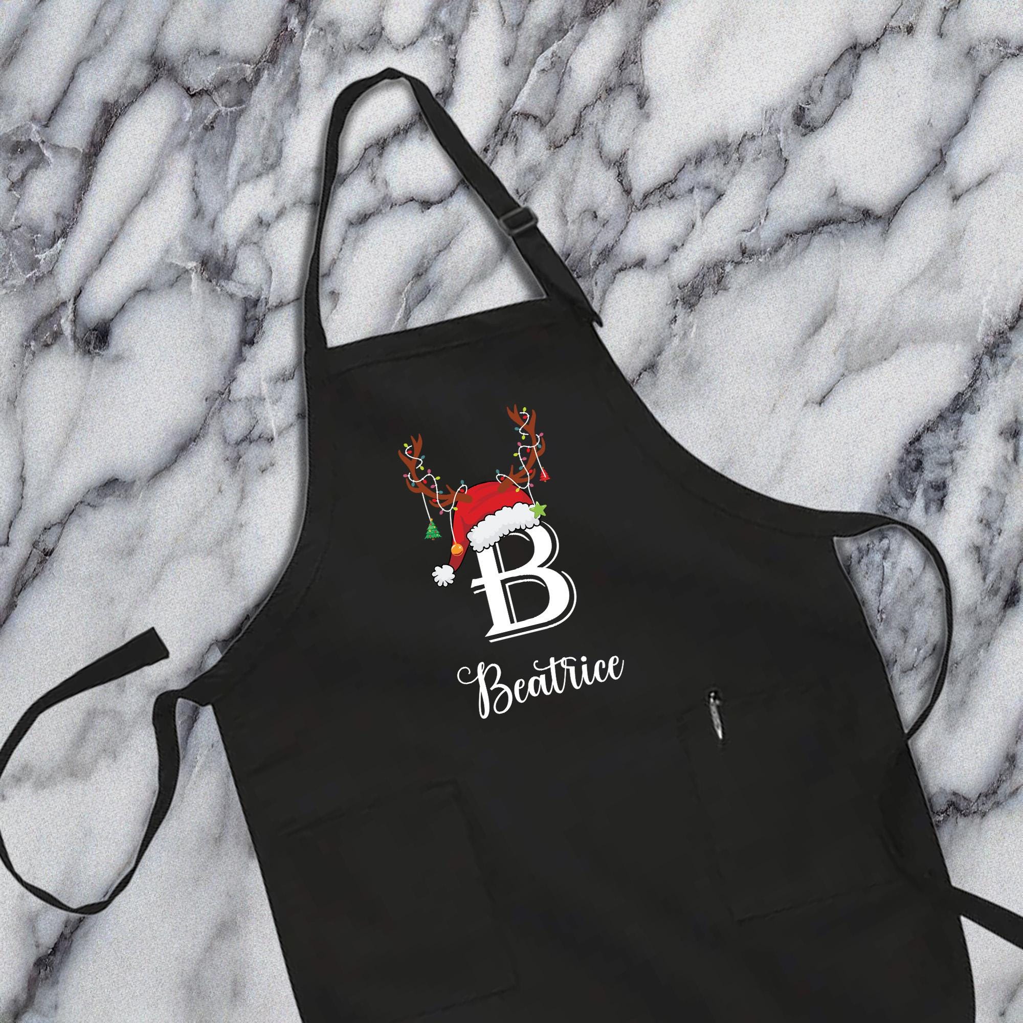 A black apron with a personalized monogrammed name and a Santa hat on it, placed on a marble surface.