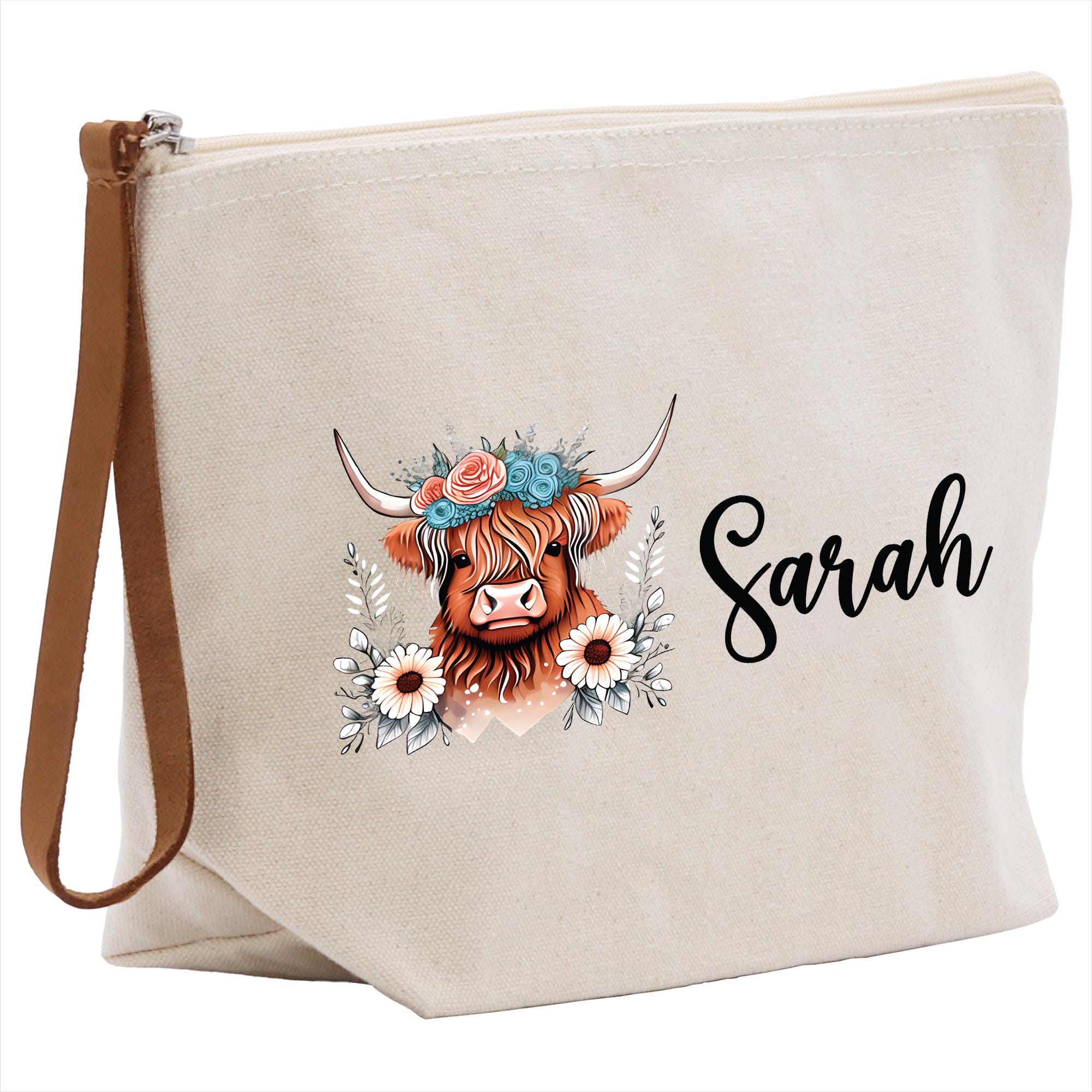 A beige canvas tote bag with a printed design of a cow&#39;s head wearing a floral crown and surrounded by flowers. The name &quot;Sara&quot; is written in cursive below the design.