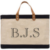 A beige tote bag with a black trim and leather handles. The bag has a monogrammed text on it that reads "B.J.S."