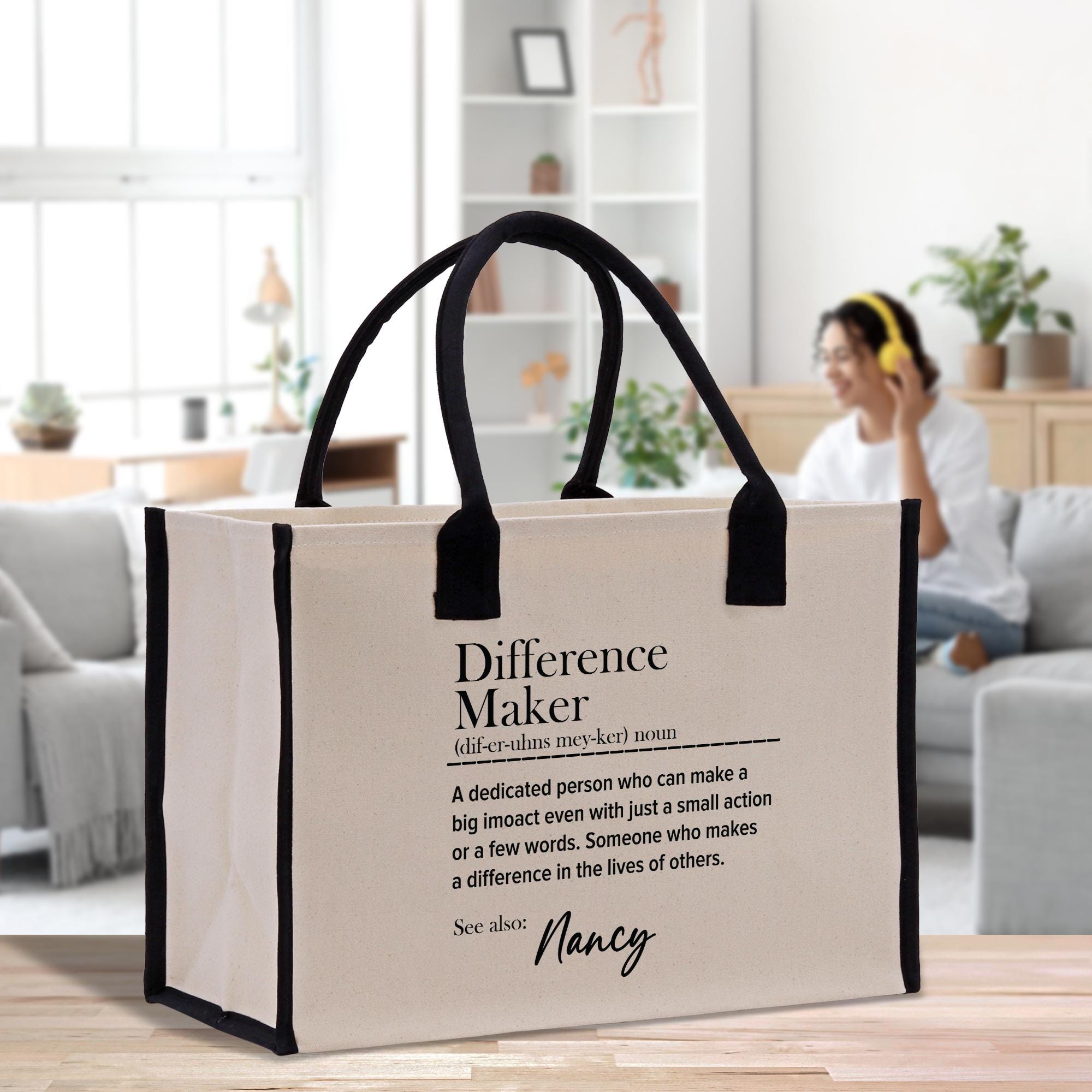 A beige tote bag with black handles, featuring a definition of a &quot; Difference Maker&quot; printed on it. The bag is placed on a wooden surface, with a person sitting on a couch in the background.
