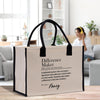 A beige tote bag with black handles, featuring a definition of a " Difference Maker" printed on it. The bag is placed on a wooden surface, with a person sitting on a couch in the background.