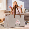 A woman is sitting on a couch in a living room, holding a phone and smiling. In front of her is a large beige tote bag with a Paris-themed design and the name "Scarflett" printed on it.