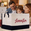 A family of three - a man, a woman, and a young girl - are gathered around a large white tote bag with the name "Jennifer" printed on it.