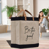 A beige tote bag with the text "Be the good" printed on it, placed on a wooden surface. In the background, there is a person sitting on a couch and reading a book.