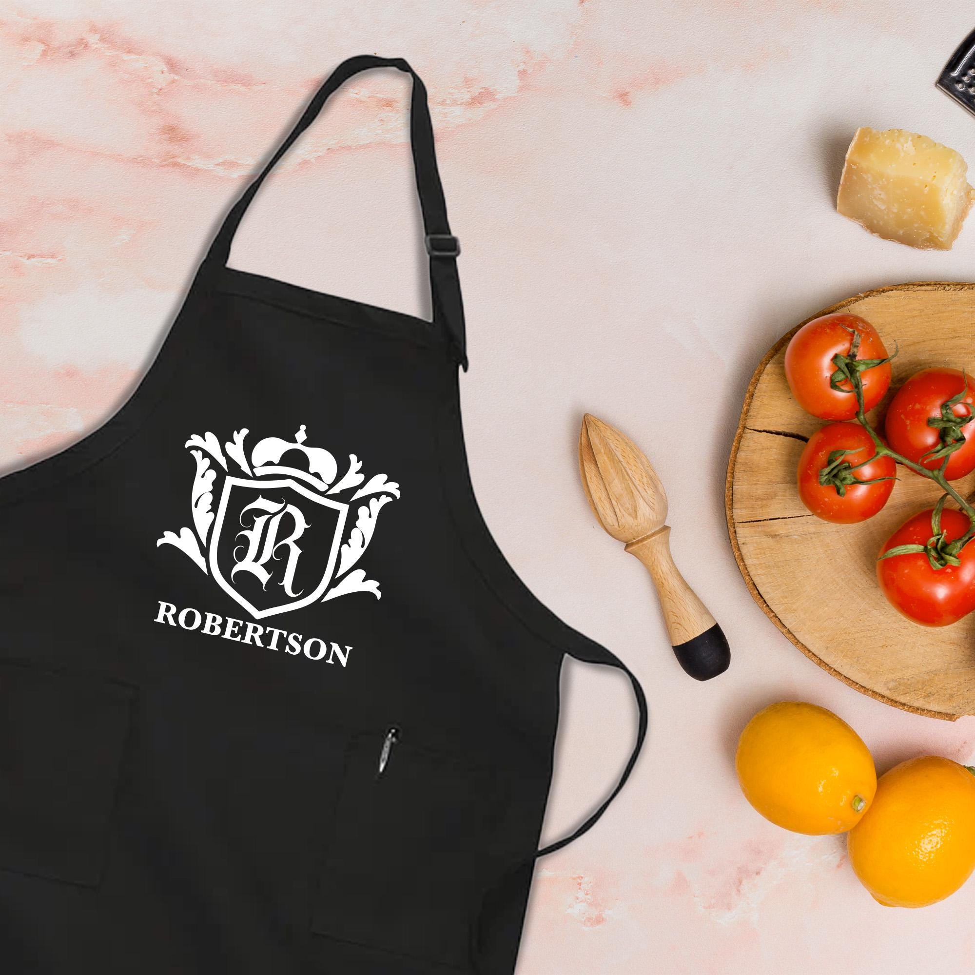 A black apron with a white coat of arms and the name &quot;ROBERTSON&quot; printed on it, along with a wooden juicer and a plate of fresh fruits, including oranges and tomatoes.