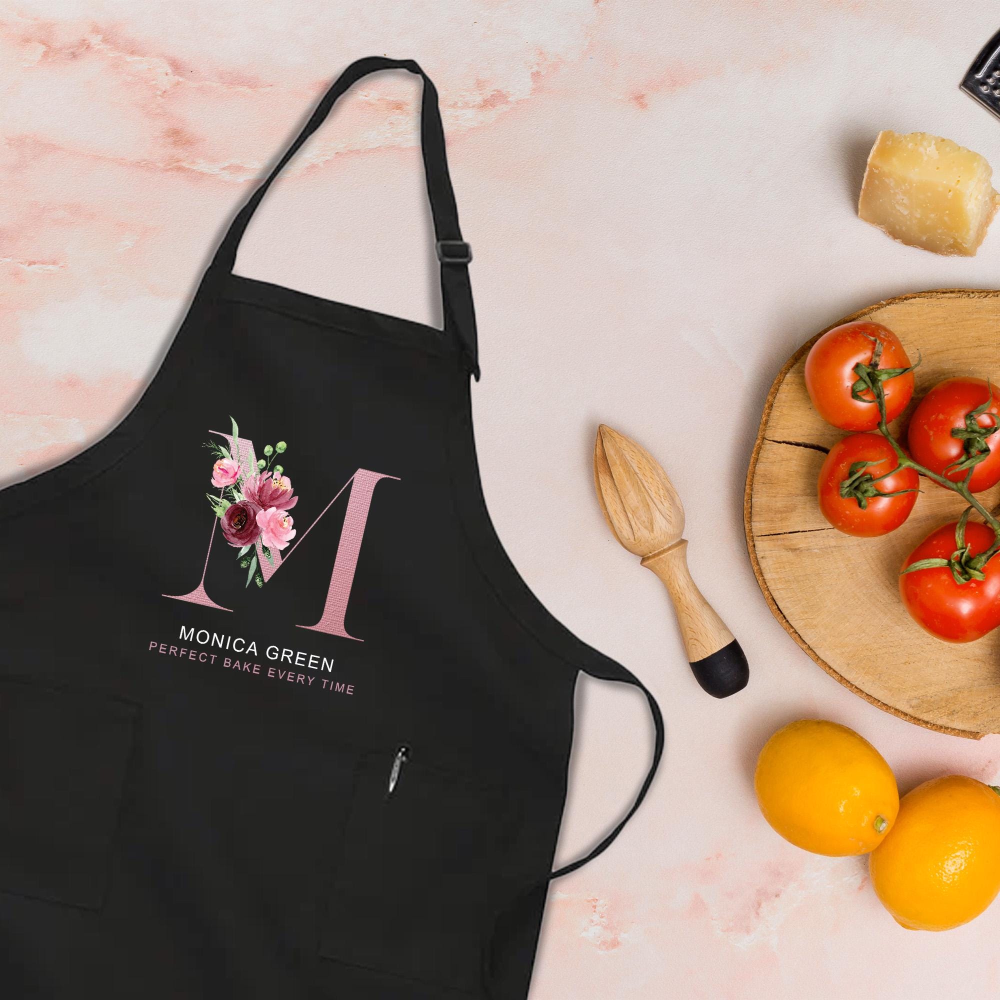 a black apron with a pink floral design, a wooden juicer, a plate of tomatoes, and some oranges on a pink marble surface.