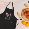 a black apron with a pink floral design, a wooden juicer, a plate of tomatoes, and some oranges on a pink marble surface.