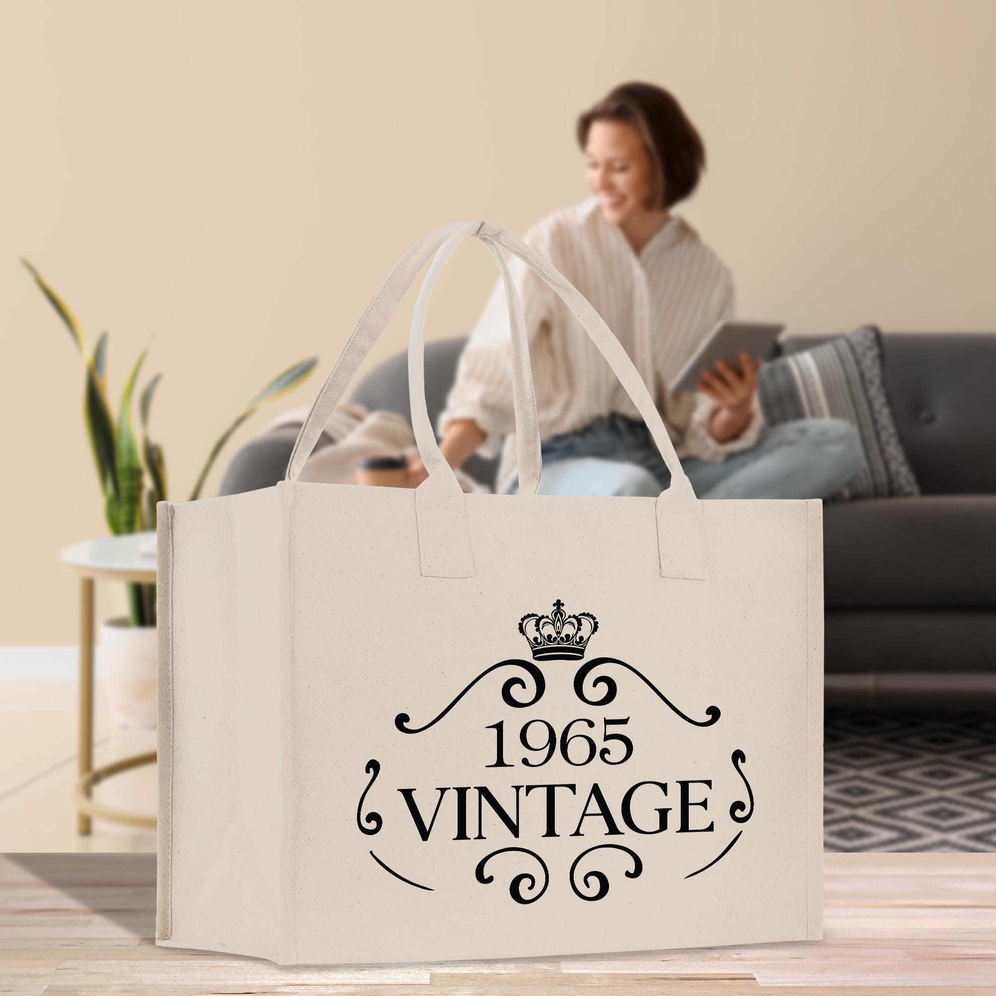 a large white tote bag with the text "1965 VINTAGE" printed on it, placed on a wooden floor in a room with a woman sitting on a couch in the background.