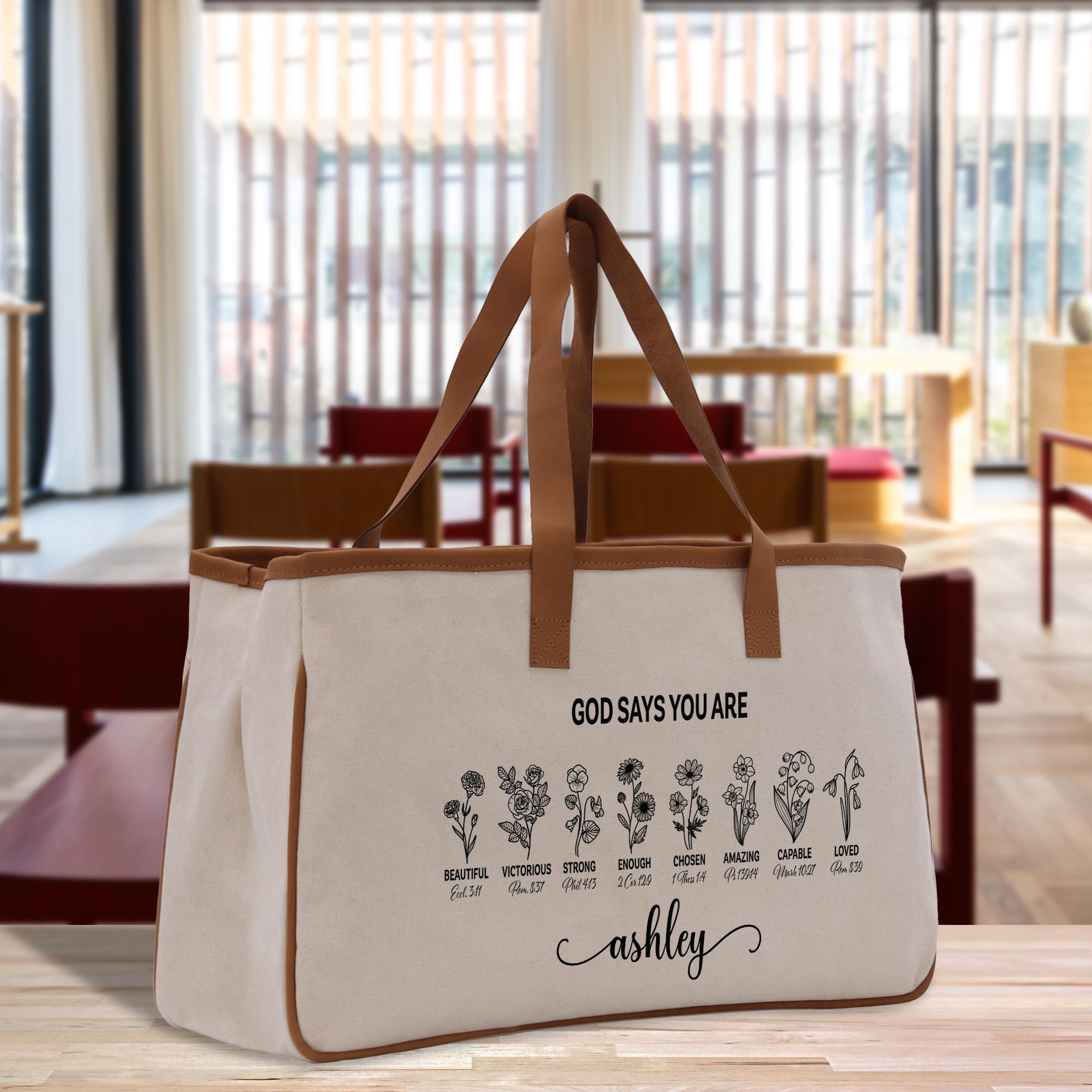 A beige canvas tote bag with a printed design of flowers and the text &quot;GOD SAYS YOU ARE Ashley&quot; printed on it, placed on a wooden surface in a room with a window.