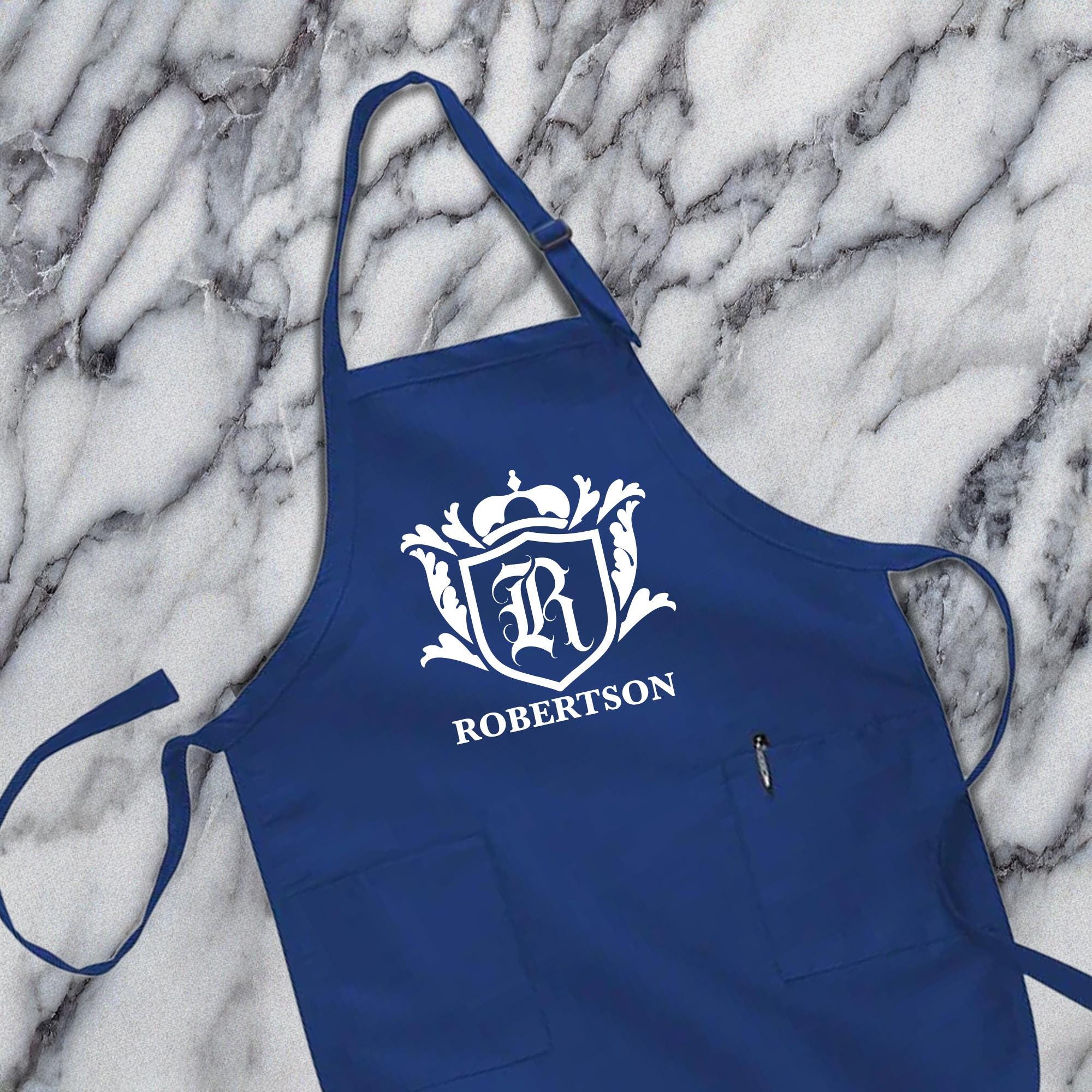 A navy blue apron with a white crest and the name &quot;ROBERTSON&quot; printed on it, placed on a marble surface.