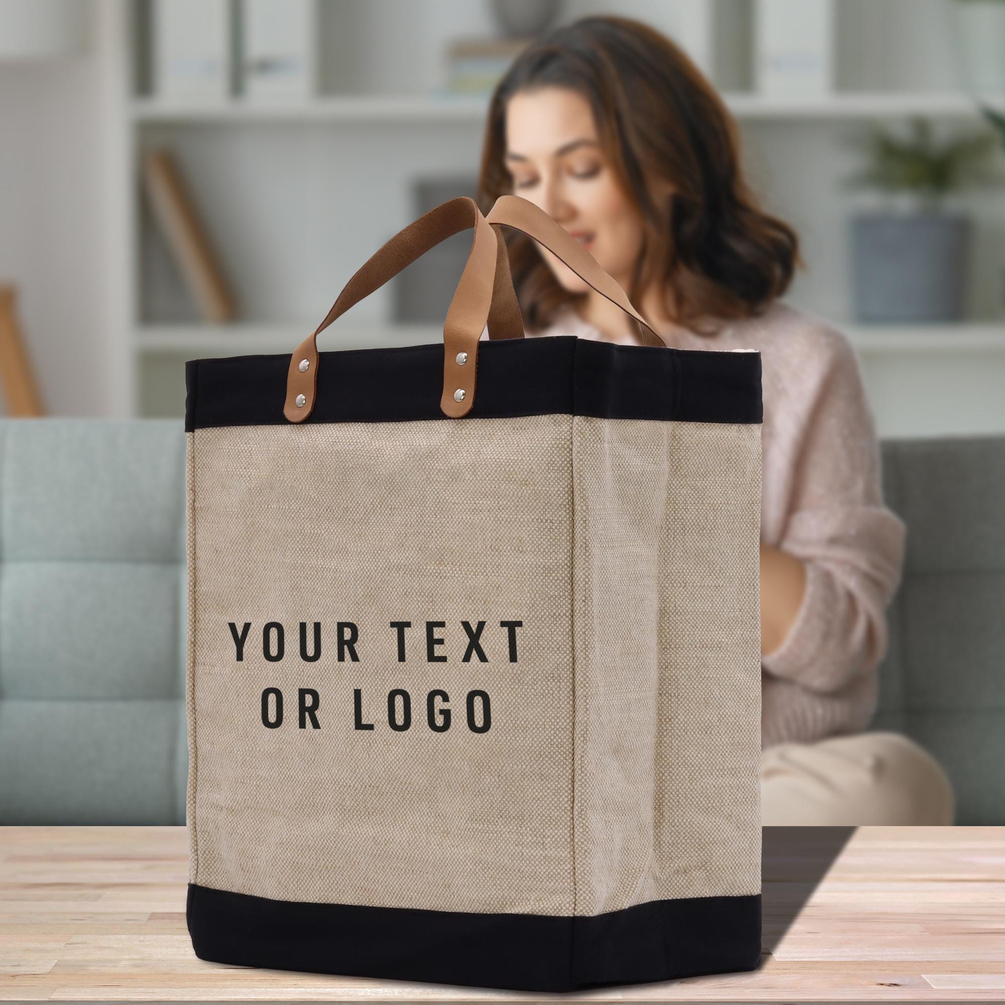 A beige tote bag with black trim and leather handles, featuring the text &quot;YOUR TEXT OR LOGO&quot; printed on it.