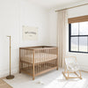 A wooden crib with a white mattress sits in a bright, white nursery room with a window and a wooden rocking chair.