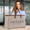 A woman is standing behind a beige tote bag with the word "NOTARY" printed on it.