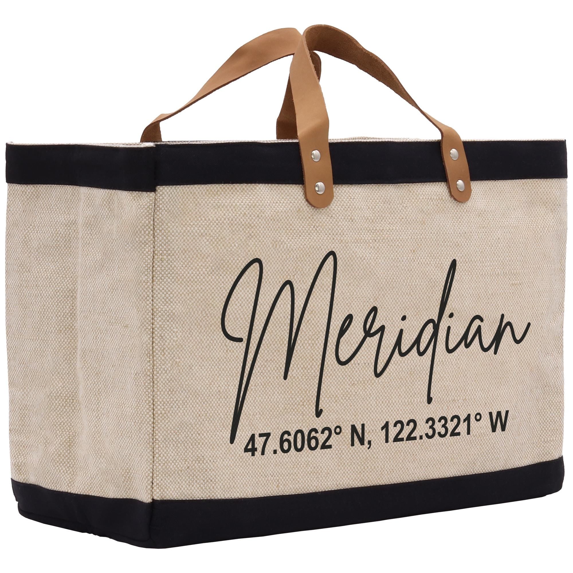 A beige tote bag with black trim and leather handles, featuring the text &quot;Meridian 47.6062 N, 122.3321 W&quot; printed on it.