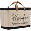 A beige tote bag with black trim and leather handles, featuring the text "Meridian 47.6062 N, 122.3321 W" printed on it.