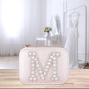 A pink makeup bag with a large letter "M" on it, placed on a wooden surface. In the background, there is a white dress and a mirror, suggesting a dressing room or dressing room setting.