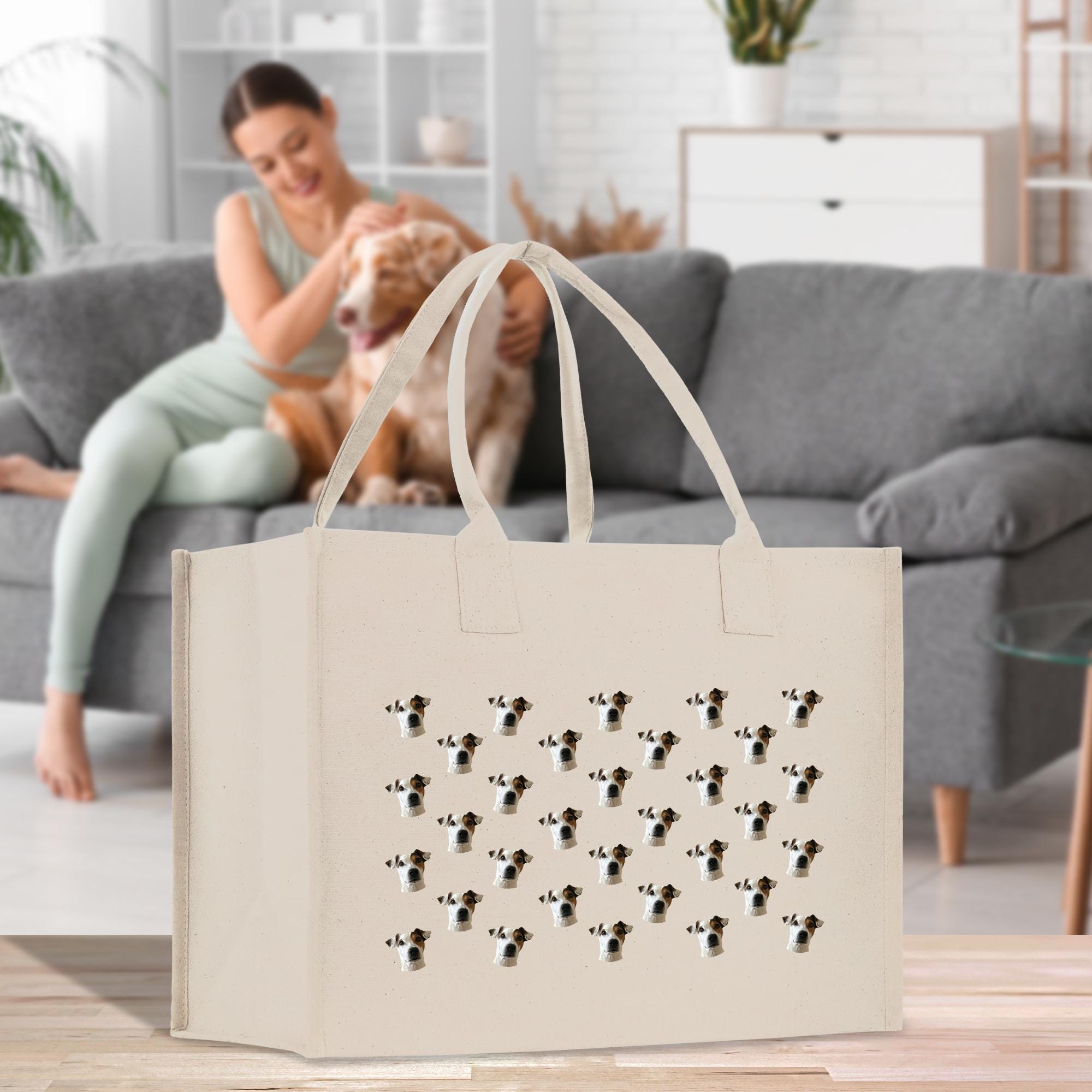 A woman is sitting on a couch in a living room, petting a dog, while a large beige tote bag with a cow pattern is placed in front of her.