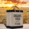 A beige tote bag with black straps and handles, featuring the text "ORGANIC FOOD just kicking IT'S WINE" printed on it, placed on a wooden surface against a backdrop of a golden sunset over a field of golden wheat.
