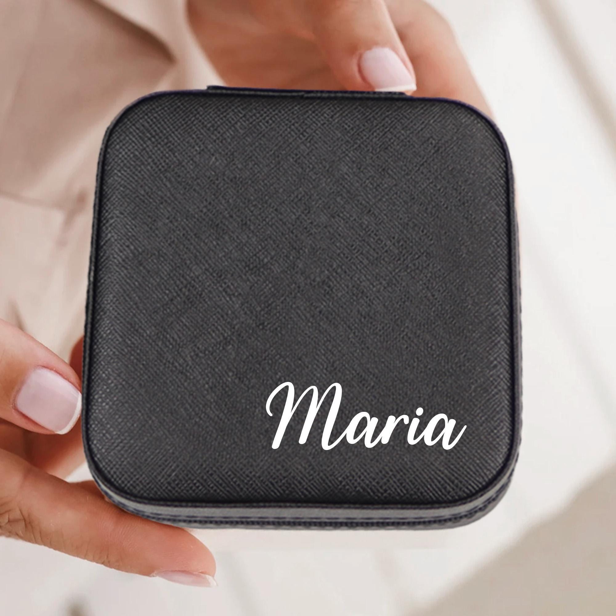 A person&#39;s hand holding a black, square-shaped case with the name &quot;Maria&quot; written on it in white text.