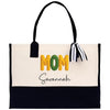 A beige tote bag with the word "MOM" printed in large letters, along with the name "Savannah" written below it. The bag has a black handle and a black and white striped ribbon tied around it.