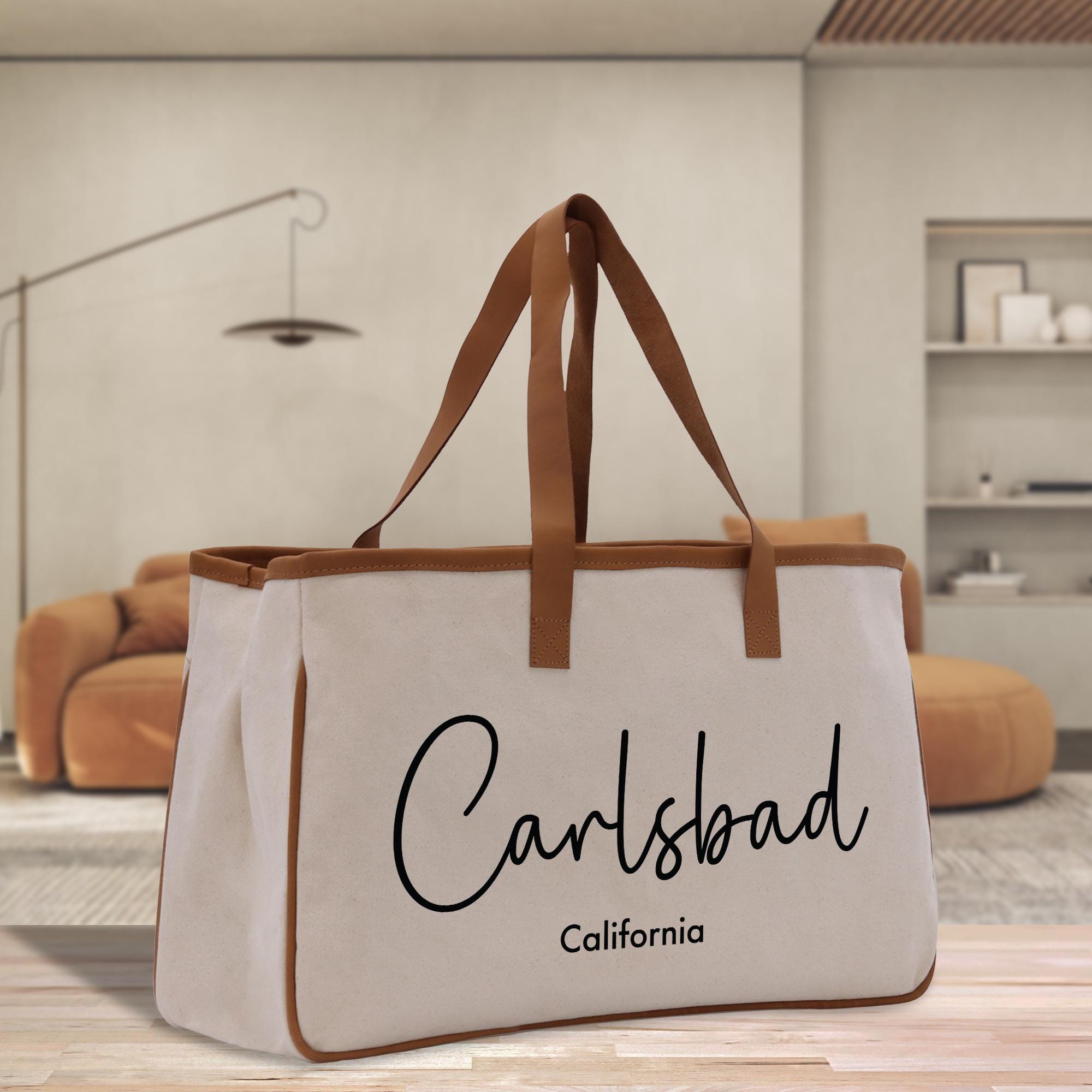 A beige tote bag with the text &quot;Carlsbad California&quot; printed on it, placed on a wooden surface in a room with a brown couch and a lamp in the background.