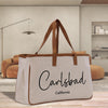 A beige tote bag with the text "Carlsbad California" printed on it, placed on a wooden surface in a room with a brown couch and a lamp in the background.