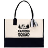 A beige canvas tote bag with a black handle and a black and white striped ribbon attached to it. The bag has a graphic of a tent and trees printed on it, along with the text 