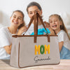 A beige tote bag with the word "MOM" printed on it, along with the name "Savannah" written below it. The bag is placed in front of three smiling women, who appear to be a mother and her two daughters.