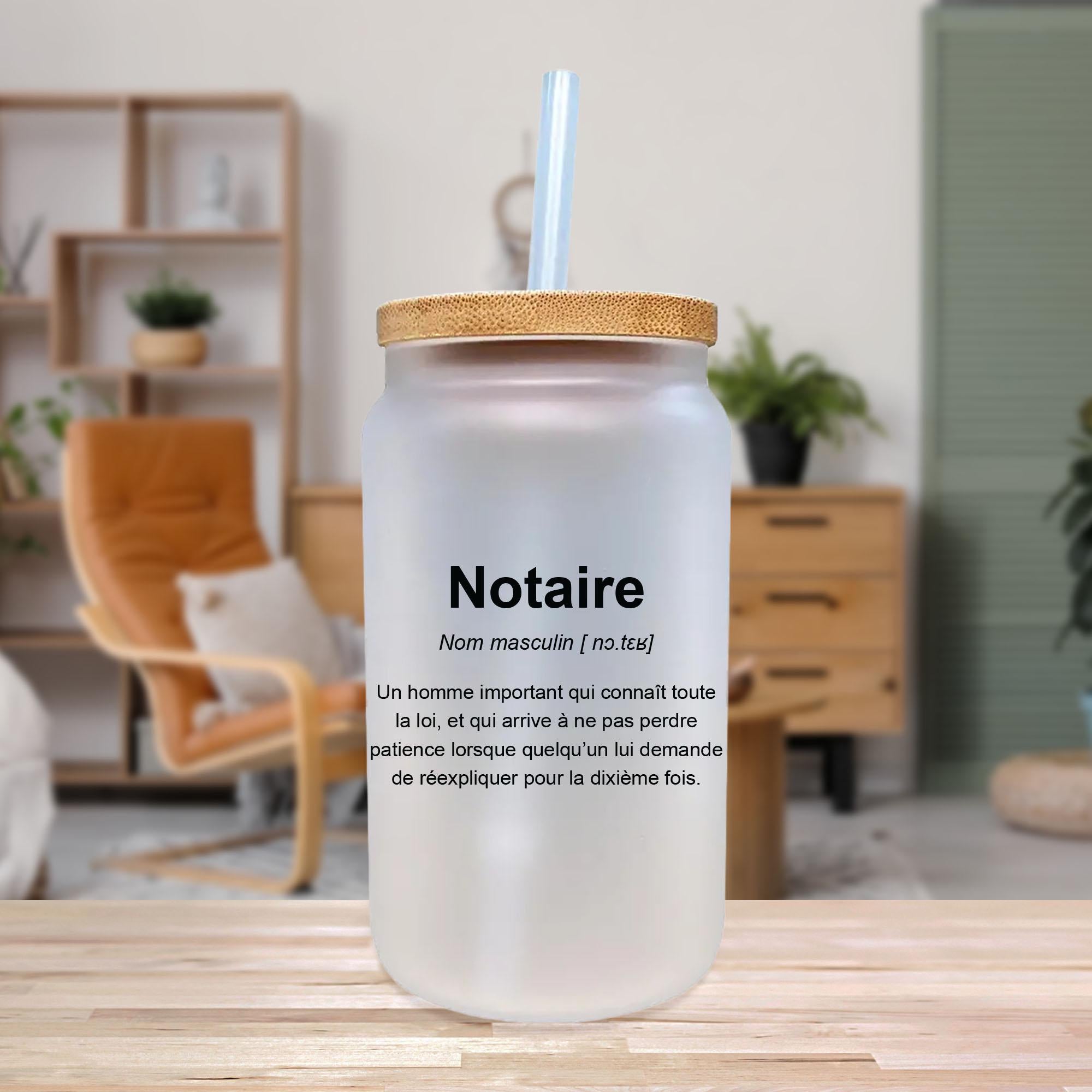 A clear glass jar with a straw, placed on a wooden surface in a room with a bookshelf and plants in the background.