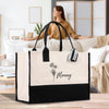 A large tote bag with a floral design and the name "Manny" printed on it, placed on a wooden surface. In the background, there is a blurred image of a woman sitting on a couch.