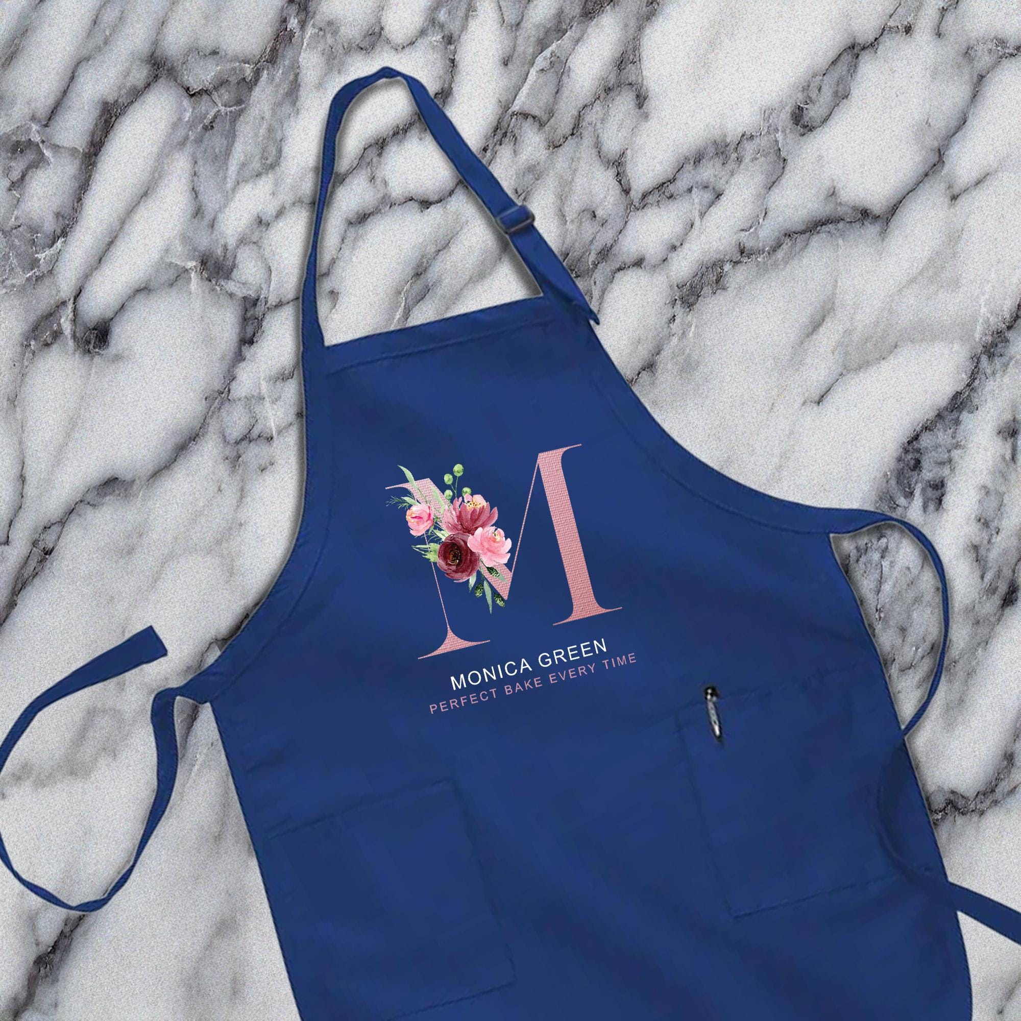 A blue apron with a floral design and the text &quot;MONICA GREEN&quot; printed on it, placed on a marble surface.