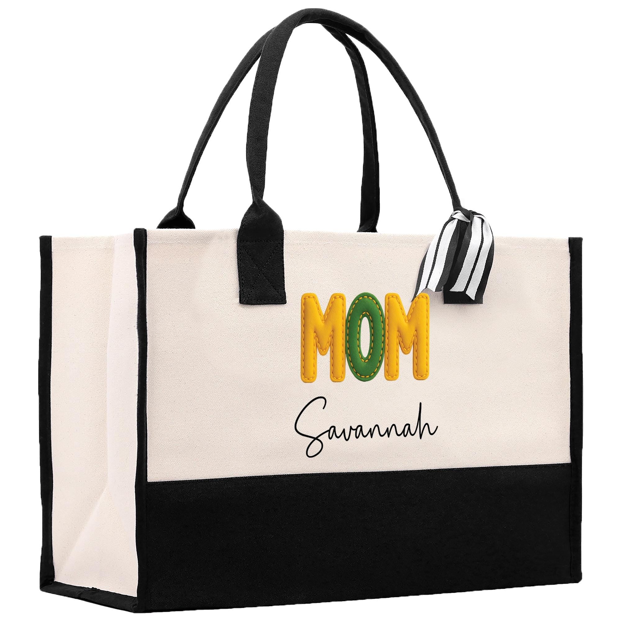 A beige tote bag with a black handle and a black and white striped ribbon attached to it. The bag has a personalized message printed on it, which reads &quot;MOM Savannah&quot;.