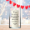 A white candle with a message written on it, placed in a glass holder on a wooden surface. The background features a blurred image of red hearts.