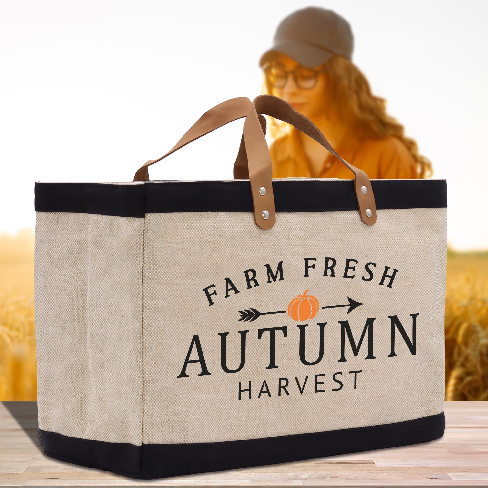 A beige tote bag with the text &quot;FARM FRESH AUTUMN HARVEST&quot; printed on it, along with an orange pumpkin symbol, is displayed on a wooden surface. In the background, there is a blurred image of a person wearing a hat and glasses.