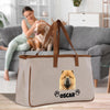 A woman is sitting on a couch, petting a dog, while a large bag with a dog image and the name "OSCAR" printed on it is placed on a wooden floor in the foreground.